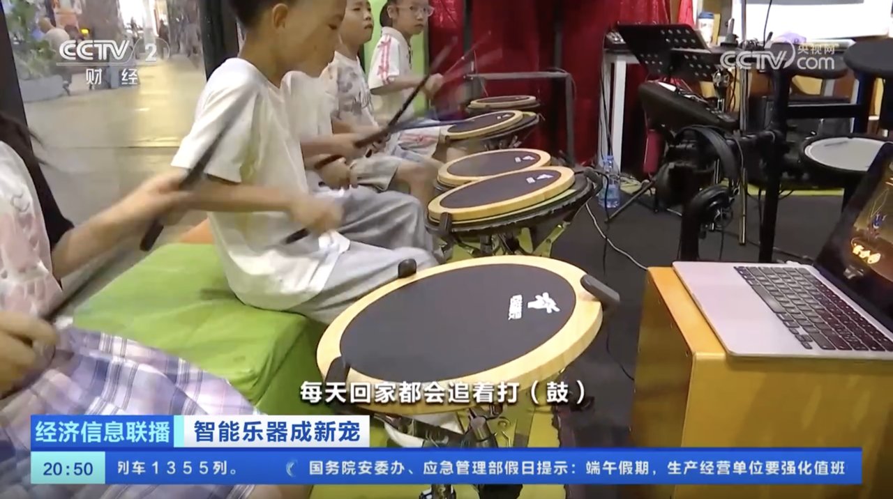 央视镜头：一排孩子坐着用诺艾哑鼓精灵练鼓，画面字幕显示每天回家都会追着打